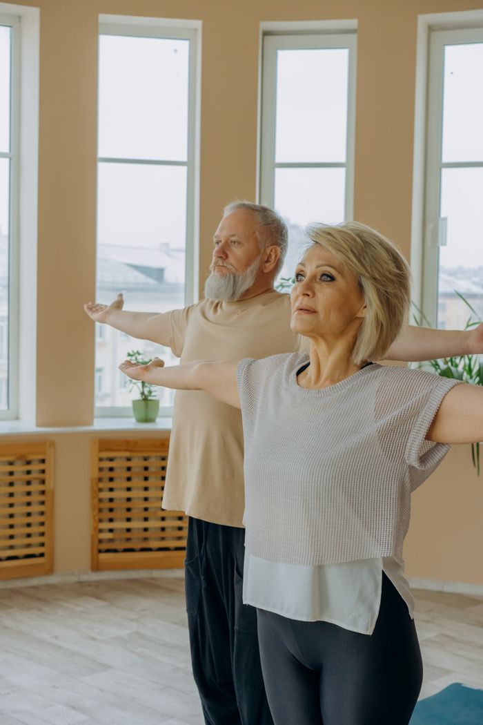 Elderly couple doing yoga indoors with arms raised, promoting health and wellness.